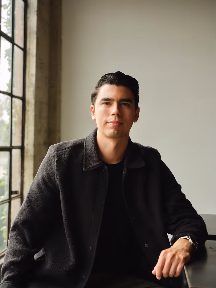 Young man with short dark hair wearing a dark jacket, sitting by a window with a neutral expression.