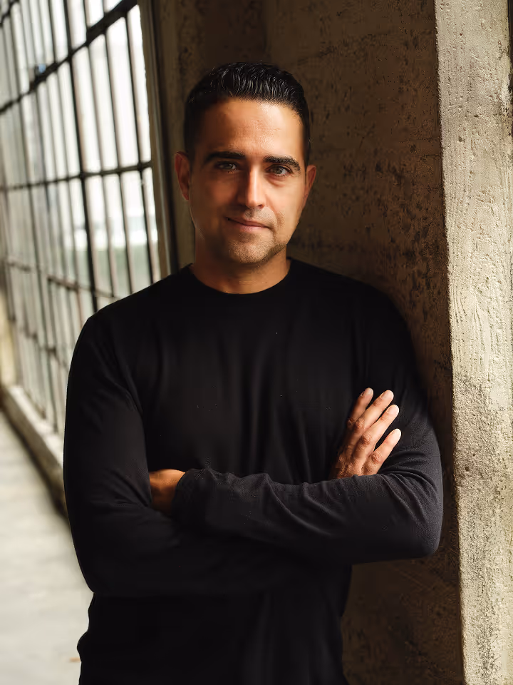 Man with short dark hair and black shirt stands with arms crossed by a textured wall near tall windows.