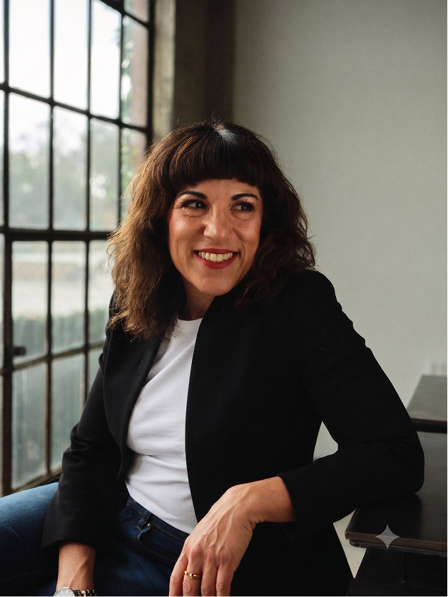Smiling woman with brown hair and bangs sitting near a window wearing a black jacket and white shirt.