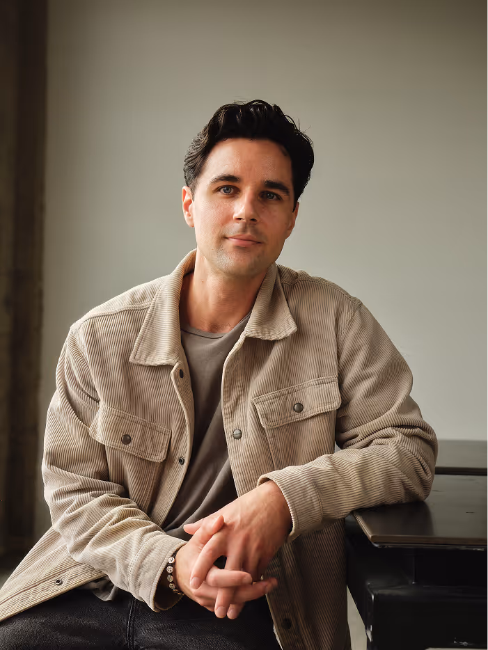 Man with dark hair wearing beige jacket and gray shirt sitting at a table with hands clasped.