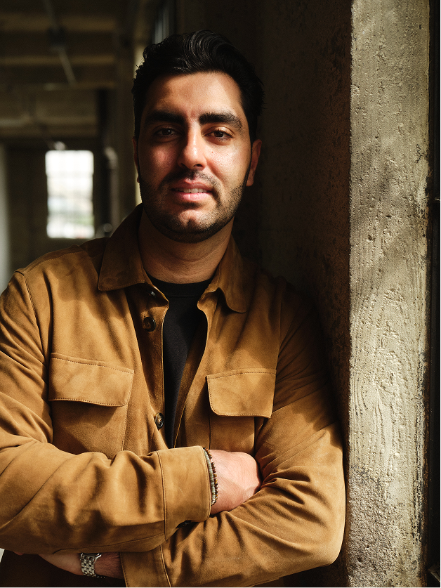 Man with dark hair and beard wearing brown jacket leaning against a concrete wall near a window.