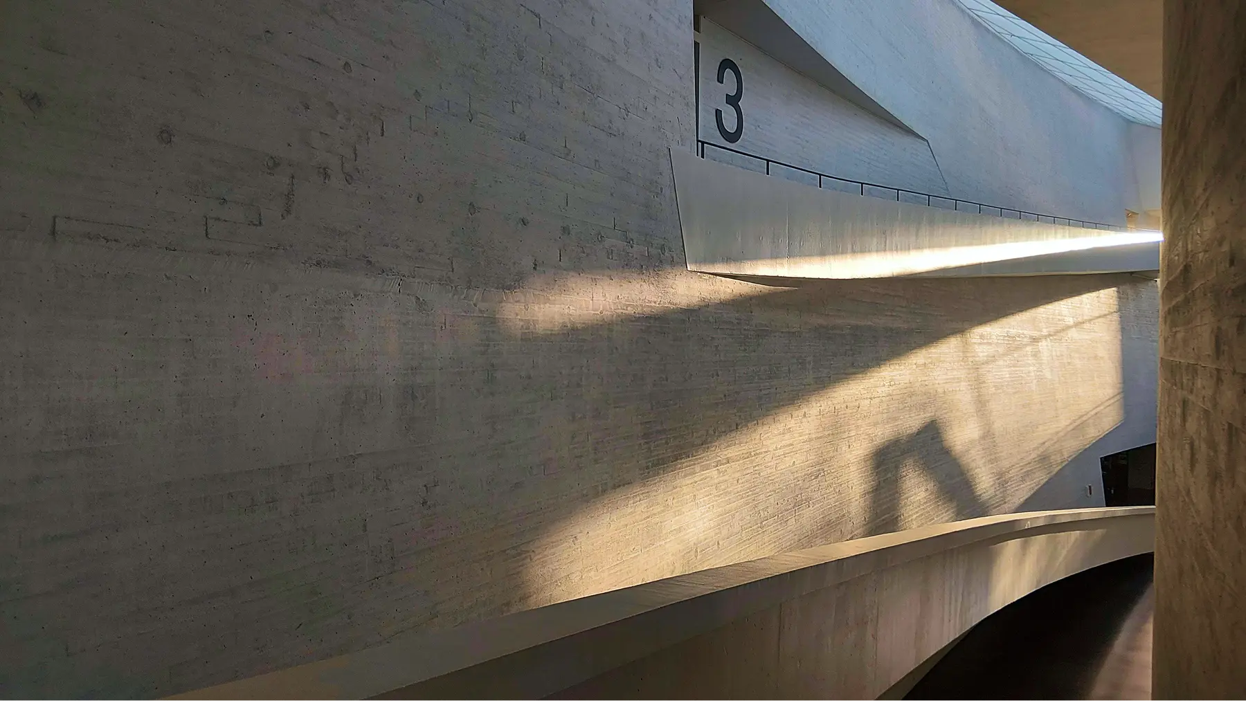 Curved concrete hallway with sunlight casting angular shadows and number 3 on the wall.