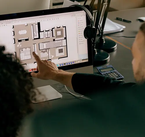 Person pointing at home floor plan displayed on a desktop computer screen in an office setting.