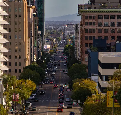 Traffic-filled urban street lined with tall buildings and trees under a clear sky.
